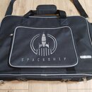 Černá přepravní taška s šedým lemováním a dvěma rukojtěmi. Na přední kapse je šedý nápis SPACESHIP a grafika rakety v kruhu. V pravé dolní části je b…