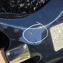Detail zadní strany černé kytary s kovovým krytem elektroniky. Na lakovaném povrchu těla jsou viditelné škrábance, které jsou zakroužkovány bílou čar…