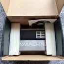 V kartonové krabici s polystyrenovými vložkami je umístěno šedé elektronické zařízení. Na přední straně je černý panel s mřížkou a nápis MACHINE. V k…
