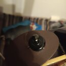 Detailní záběr na hnědý, texturovaný materiál s kulatým černým knoflíkem uprostřed. V pozadí je vidět část světlého nástroje s hřistnicí a strunami.…