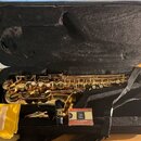 Zlatý saxofon v černém kufru. Vedle nástroje jsou položeny žlutý látkový sáček s dvěma rýdy a krabička rýd s logem „G“ a nápisem „GENUINE“. Na krabič…