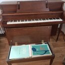 Hnědé dřevěné piano s klaviatura. Na čelní straně nad klávesami je vyryto logo WILLIS. Před nástrojem stojí otevřený dřevěný stolek s dvěma notovými…