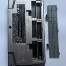 Zadní strana šedého elektronického zařízení s otevřeným úložným prostorem pro baterie a vedle něj položeným šedým plastovým víkem. Na spodní straně j…