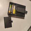 Černý plastový držák na baterie s odpojeným víkem. Uvnitř jsou tři cylindricalní baterie s nápisy GP, Panasonic 1000 a nečitelným žlutým označením. N…
