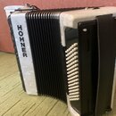 Bílá akordeon s černým měchovcem a černými detaily. Na bílém povrchu je svisle napsán nápis HOHNER. Nástroj má bílé tlačítka a černý popruh. Povrch v…