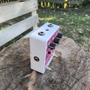 Bílý kytarový efekt pedál v kovovém šasi s červeným grafickým potiskem na horní straně. Na pravé straně jsou tři černé oválné kontrolní knoflíky a na…
