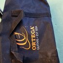 Černý textilní pouzdro na hudební nástroj s dvěma černými popruhy. Na povrchu je oranžové logo a bílý nápis ORTEGA GUITARS • EST 1994. Materiál vykaz…