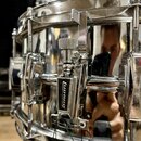 Detailní záběr na chromovaný snare drum s vysokým leskem. Na mechanismu je viditelný nápis "diman". Povrch nástroje vykazuje drobné škrábance a otřep…