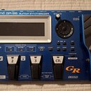 Modrý kytarový syntetizátor s nápisem Roland GR-55 GUITAR SYNTHESIZER. Zařízení má displej, ovládací knoflík se šipkami a čtyřistupňové pedály označe…