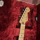 Hlava a hриф elektrické kytary z jasného dřeva v červeném plišovém kufru. Na hlavě je černý nápis Fender® STRATOCASTER a v horní části sériové číslo…