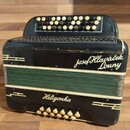 Černá harmonika s zeleným měchovcem. Na přední straně je bílý nápis Josef Hlaváček Louny a níže nápis Heligonka. Nástroj vykazuje známky opotřebení v…