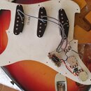 Fotografie zachycuje vnitřní část elektrické kytary s bílým pickguardem a tělem v červeno-žlutém sunburst provedení. Na bílém plastu jsou tři černé s…