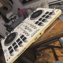 Bílý DJ 컨트롤러 s dvěma jogovými koly a černými kontrolními tlačítky. Na přístroji jsou viditelné nápisy Numark a MIXTRACK PRO II. Zařízení je umístěno…
