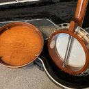 Fotografie zachycuje rozložený banjo v černém etuýru. Nástroj má hnědý dřevěný korpus a bílou blánu, na které je vidět částečně nečitelná nálepka. Ko…
