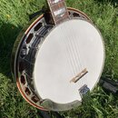 Bílá banjo s kovovým ráfkem a hředy. Na hmatníku jsou viditelné perleťové vložky ve tvaru kapek. Nástroj je umístěn na černém stojanu v travě.