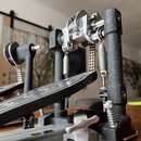 Detailní záběr na mechanismus bicího pedálu v černé a stříbrné barvě. Jsou vidět kovové řetízky, pružiny a nastavovací šrouby. Na komponentech nejsou…