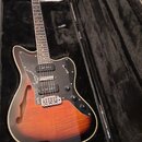 Polodupatá elektrická kytara s tělem v oranžovém sunburst provedení a černým pickguardem. Na hlavě hředu je viditelné logo Ibanez. Nástroj je umístěn…