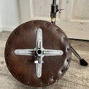 Kruhová sedací skořápě v hnědé kůži s kovovým křížovým mechanismusem uprostřed. Sedák je připevněn k podvozku pomocí viditelných šroubů po celém obvo…