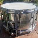Stříbrný kovový snare drum s bílou blánou. Na povrchu korpusu jsou viditelné škrábance a známky opotřebení. Nástroj je vybaven chromovaným obručování…