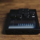 Černý elektronický kontrolér s displejem a několika ovládacími tlačítky v horní části. Na spodní hraně jsou viditelné konektory s nápisy MIDI a AUDIO…