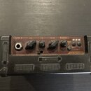 Kytarový kombo zesilovač s černým krytím a hnědým ovládacím panelem. Na panelu jsou nápisy GUITAR IN, GAIN, TONE, VOLUME a RHYTHM FX, dále přepínač M…