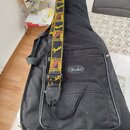 Černý polstrovatý obal na kytaru s nápisem Fender v oválném logu. Na obalu leží kytarový popruh s žlutým nápisem Fender a hnědo-žlutými grafickými pr…