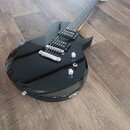 Černá elektrická kytara s tvarem typu Les Paul a lesklým povrchem. Nástroj má dva humbuckery, dvě kontrolní knoflíky a jeden přepínač. Na kytaru nejs…