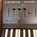 Černý klavír s dřevěnými bočními panely. Na horním panelu jsou bílé nápisy CRUMAR a PERFORMER, dále označení BRASS a FILTER CONTROLS. Jsou zde posuvn…