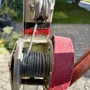 Fotografie zachicuje detail mechanismu s ocelovým lanim namotaným na červenou cívku. Součástí je kovová kladka a vodící lišta v červeno-černé barvě.…