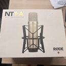 Bílá krabice s nápisem NT1-A STUDIO CONDENSER MICROPHONE a logem RØDE MICROPHONES. Uprostřed je znázorněn stříbrný kondenzátorový mikrofon v černém s…