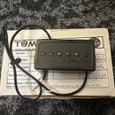 Černý humbucker s šesti viditelnými šrouby v polosovém uspořádání. Na povrchu je rukopisní nápis Tuma. Snímač má připojený černý kabel a leží na bílé…