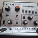 Stříbrný elektronický nástroj s nápisem Stylophone THEREMIN. Zařízení obsahuje několik otočných knoflíků, přepínačů a mřížku reproduktoru. Na panelu…