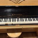 Dřevěný piano s černým lakovaným krytem klaviatury, na kterém je zlatý nápis A.PROKSCH. Klaviatura vykazuje známky opotřebení, zejména u několika bíl…