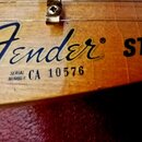 Detail dřevěné hlavy nástroje s nápisem Fender a textem SERIAL NUMBER CA 10576. Vedle loga je vidět začátek slova STR. Povrch dřeva vykazuje drobné z…