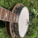 Banjo s bílou blánou a kovovým kroužkem kolem těla. Hmatník je z tmavého dřeva s vloženými světlými ozdobnými značkami v podobě kapek. Na těle nástro…