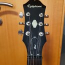 Černá hlava kytary s bílým nápisem Epiphone v horní části a nápisem Dot na spodní části. Uprostřed se nachází bílý ozdobný vložkový prvek. Jsou zde v…