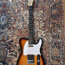 Elektrická kytara typu Telecaster s tělem v provedení sunburst a bílým pickguardem. Na hlavě hředu je viditelné logo "Fender". Kytara má tmavý hmatní…