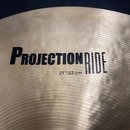 Detailní záběr na povrch mosazného činely s koncentrickým žebrováním. Na povrchu je černý nápis PROJECTION RIDE a pod ním menší text 21"/53 cm. Povrc…