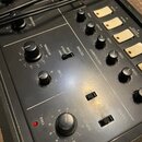 Detailní pohled na ovládací panel šedého elektronického zařízení s několika černými otočnými knoflíky a bílými tlačítky. Na panelu jsou viditelné náp…