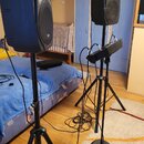 Dvě černé aktivní reproduktory umístěné na černých stativech. Na mřížce pravého reproduktoru je viditelné malé logo, které je však nečitelné. Mezi re…