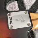 Detail zadní strany kytary s kovovým krytem elektroniky připevněným čtyřmi šrouby. Na krytu je vyryto logo ve tvaru stylizované čtverné sedmičky a ná…