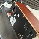 Zadní strana reproduktoru s dřevěným povrchem a černým zadním panelem. Na panelu je vypínač s nápisy POWER, ON a OFF, vstup AC IN s připojeným kabele…