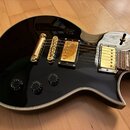 Černá elektrická kytara s lesklým povrchem a bílým lemováním hran. Nástroj má zlatý hardware, včetně dvou humbuckerů a mostu. Na těle jsou čtyři čern…