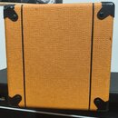 Fotografie zachycuje zadní stranu kytarového zesilovače potaženého oranžovým texturovaným vinylem. V rozích jsou umístěny černé plastové rohové chrán…