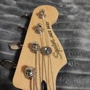 Fotografie zachicuje hlavu basové kytary z přírodního světlého dřeva s chromovanými ladičkami. Na hlavě je černý nápis Squier JAZZ BASS a pod ním men…