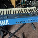 Modrý synthesizér s klaviatura umístěný na černém stojanu. Na přední straně je bílý nápis YAMAHA a vpravo označení modelu CS1x. Na ovládacím panelu j…