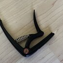 Černý kytarový kapotastán z kovového materiálu s pružinou. V místě spoje je vidět malý červený kruhový prvek. Na předmětu nejsou žádné čitelné značky…