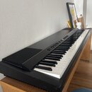 Černý digitální klavír s bílými a černými klávesami, umístěný na dřevěném stole. Na horním ovládacím panelu jsou vidět tlačítka a přepínače, ale žádn…