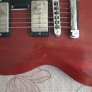 Detail korpusu červené elektrické kytary s dvěma humbuckery a chromovaným mostem. Na povrchu laku jsou viditelné škrábance a oděrky v oblasti mezi sn…