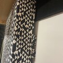 Fotografie zachycuje detail akustického panelu potaženého látkou s černobílým geometrickým vzorem tvořeným trojúhelníky. Panel je umístěn vedle bílé…